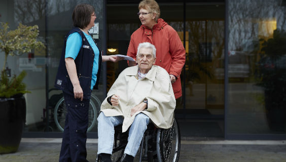 En sosu-assistent står foran et plejecenter sammen med en frivillig og en beboer. Beboeren sidder i kørestol og venter på, at den frivillige tager ham til lægen. 